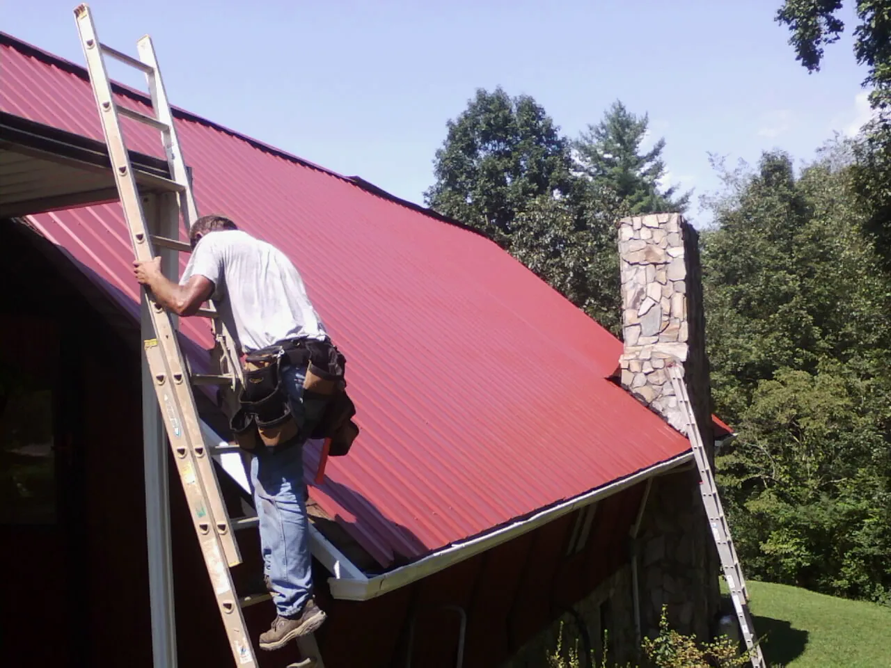 Roofer on a ladder beside a red metal roof and stone chimney — Soffit & Fascia Repair by Roofing Brothers in Whitefield