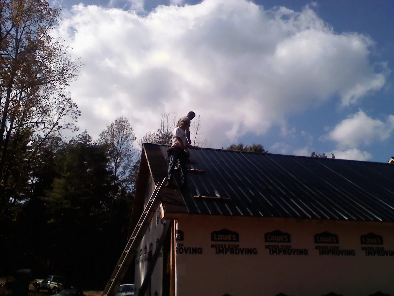 Roofer fastening dark metal panels high against a cloudy sky, from a Roof Inspection job in Whitefield